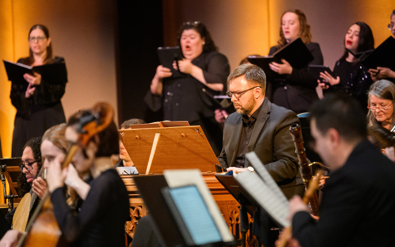 Four singers and orchestra members, surrounding organ performer