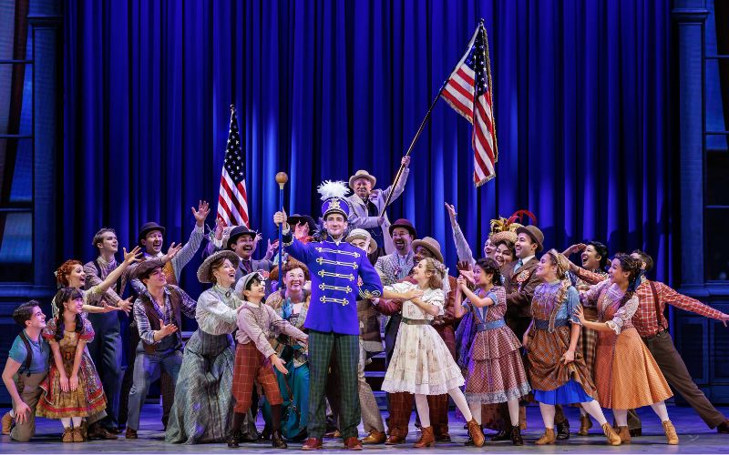 Cast members celebrate around Harold Hill during The Music Man beneath American flags.
