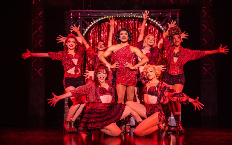 The cast of Kinky Boots performs a high-energy number in coordinated red costumes under vibrant stage lighting.
