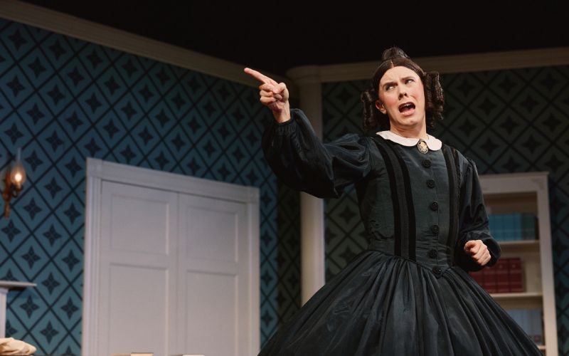 A person dressed in a mid-1800's black gown with brown hair and curls by their face pointing a finger dramatically at something off camera.
